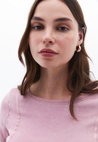 Rosa Baumwollbluse mit Rundhalsausschnitt und sichtbaren Nahtdetails; das Modell trägt große goldene Creolen; glatte Textur, natürliche Oberfläche.