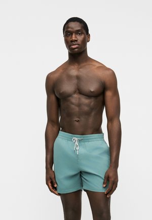 Homme à la carrure musclée, debout face à l’avant, portant un short de bain bleu clair avec un cordon blanc sur un fond uni.