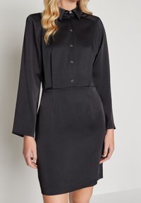 Svart skjortklänning med långa ärmar, gjord av ett mjukt tyg. Har en krage, veckdetaljer fram och en figurnära silhuett.