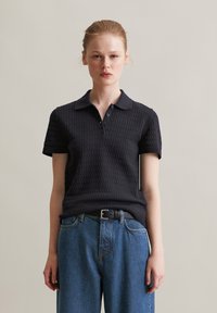 Zwarte poloshirt met textuurstof, korte mouwen en een kraag. Gecombineerd met high-waisted blauwe denim jeans en een zwarte riem.