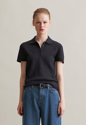 Zwarte poloshirt met textuurstof, korte mouwen en een kraag. Gecombineerd met high-waisted blauwe denim jeans en een zwarte riem.