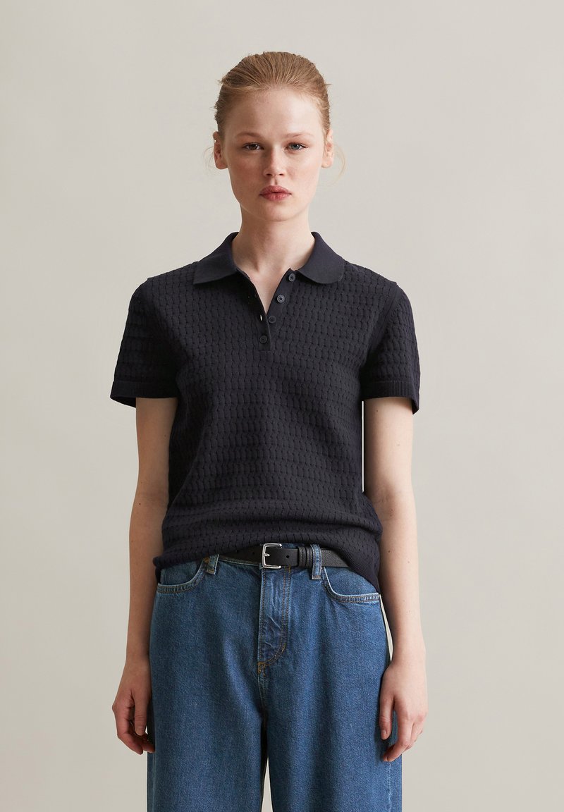 Zwarte poloshirt met textuurstof, korte mouwen en een kraag. Gecombineerd met high-waisted blauwe denim jeans en een zwarte riem.