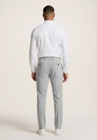 Mand, der står med ryggen til, iført en hvid langærmet skjorte, lysegrå bukser med baglommer og hvide sneakers på en ensfarvet baggrund.