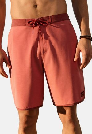 Man wearing coral-colored swim shorts with dark red waistband and drawstring, standing with hands relaxed at sides against white background.