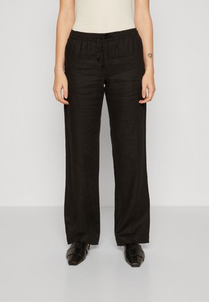 Vrouw draagt zwarte, wijd uitlopende broek met elastische tailleband en zwarte schoenen met vierkante neus, staand tegen een effen lichte achtergrond.