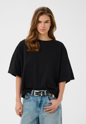 Kvinne med bølgete brunt hår har på seg en oversized svart T-skjorte stukket inn i lyseblå jeans med svart belte, står foran ensfarget bakgrunn.