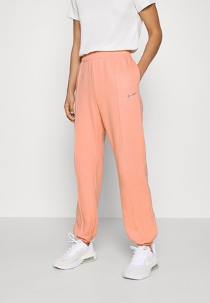 Personne portant un pantalon de survêtement Nike couleur pêche, des baskets blanches et un t-shirt blanc à manches courtes, debout devant un fond uni.