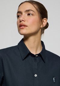 Camicia blu navy con texture uniforme, colletto classico e un piccolo logo ricamato. Bottone bianco al colletto e alla placca.