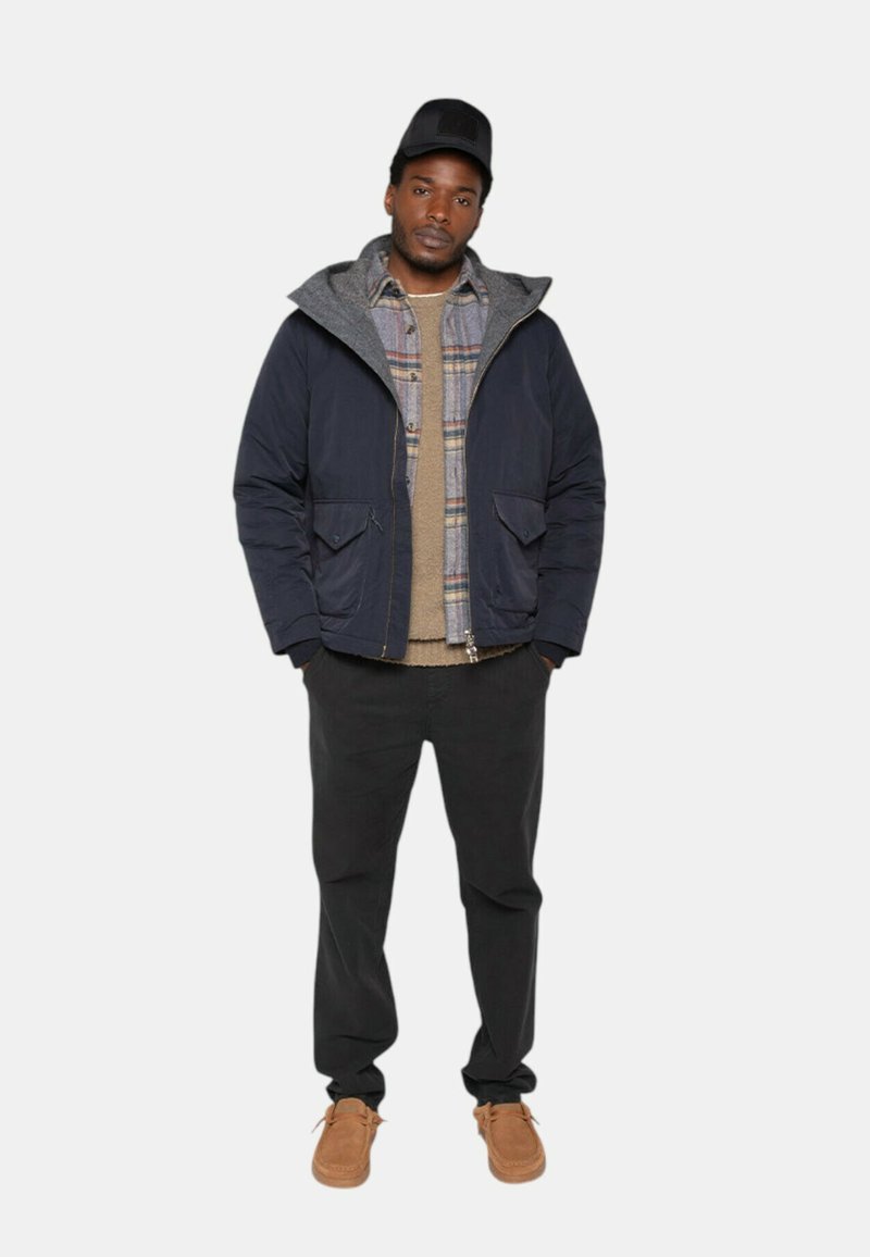 Veste bleu marine avec capuche grise, chemise à carreaux texturée en dessous, pull beige, pantalon sombre et chaussures brunes. Une casquette simple complète la tenue.