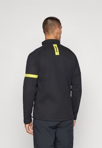 Pull-over technique noir avec col montant, texture côtelée et accents jaunes au niveau du col arrière et des manches, conçu pour le sport.