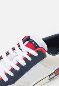 Sneaker bianca e blu navy con dettagli rossi, materiali in pelle e suede, design classico con lacci, dotata di un'etichetta marchiata sulla linguetta.