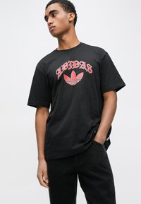 Melna kokvilnas T-krekls ar sarkanu Adidas logotipu un trim svītrām virs ziedu grafikas, brīvs piegriezums, īsas piedurknes un apaļa kakla izgriezums.