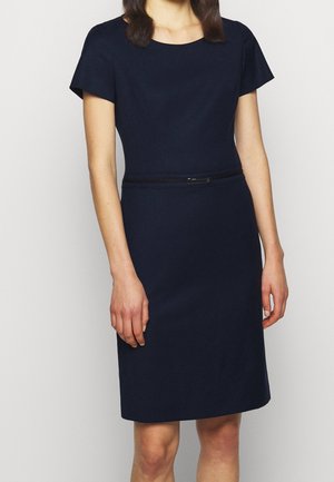 Kobieta ubrana w dopasowaną, granatową sukienkę do kolan z krótkimi rękawami i wąskim paskiem w talii, stojąca na jednolitym tle.