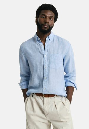 Light blue linen button-up shirt with long sleeves and single chest pocket, paired with cream-colored pleated trousers and a brown belt.