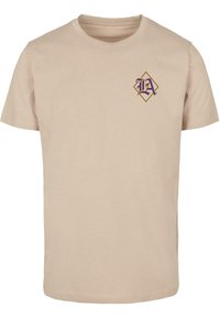 Camiseta de algodón beige con mangas cortas, que presenta un logo de diamante púrpura y dorado en el área del pecho izquierdo. Diseño simple de cuello redondo.