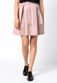 Ljust rosa plisserad kjol med hög midja och slät textur, i kombination med svarta lackskor med snörning. Kjolen har en utsvängd silhuett.