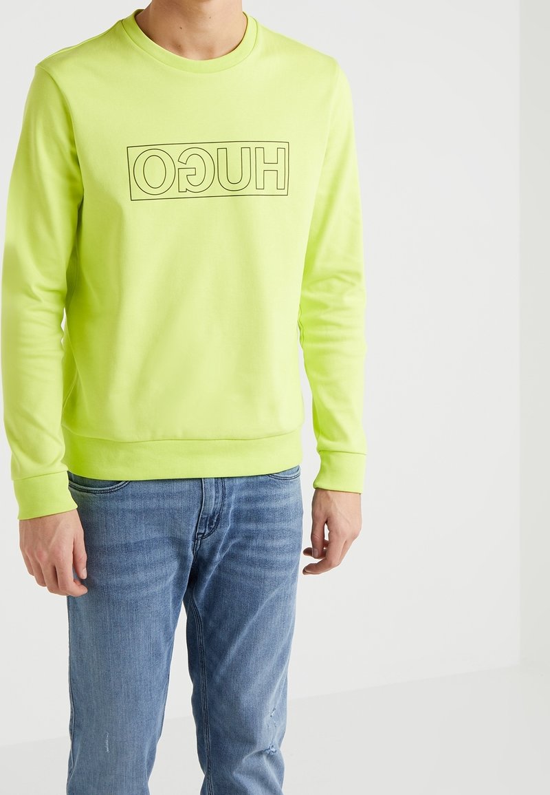 Personne portant un sweat-shirt jaune-vert vif avec un texte "HUGO" inversé et un jean bleu, debout devant un fond blanc uni.