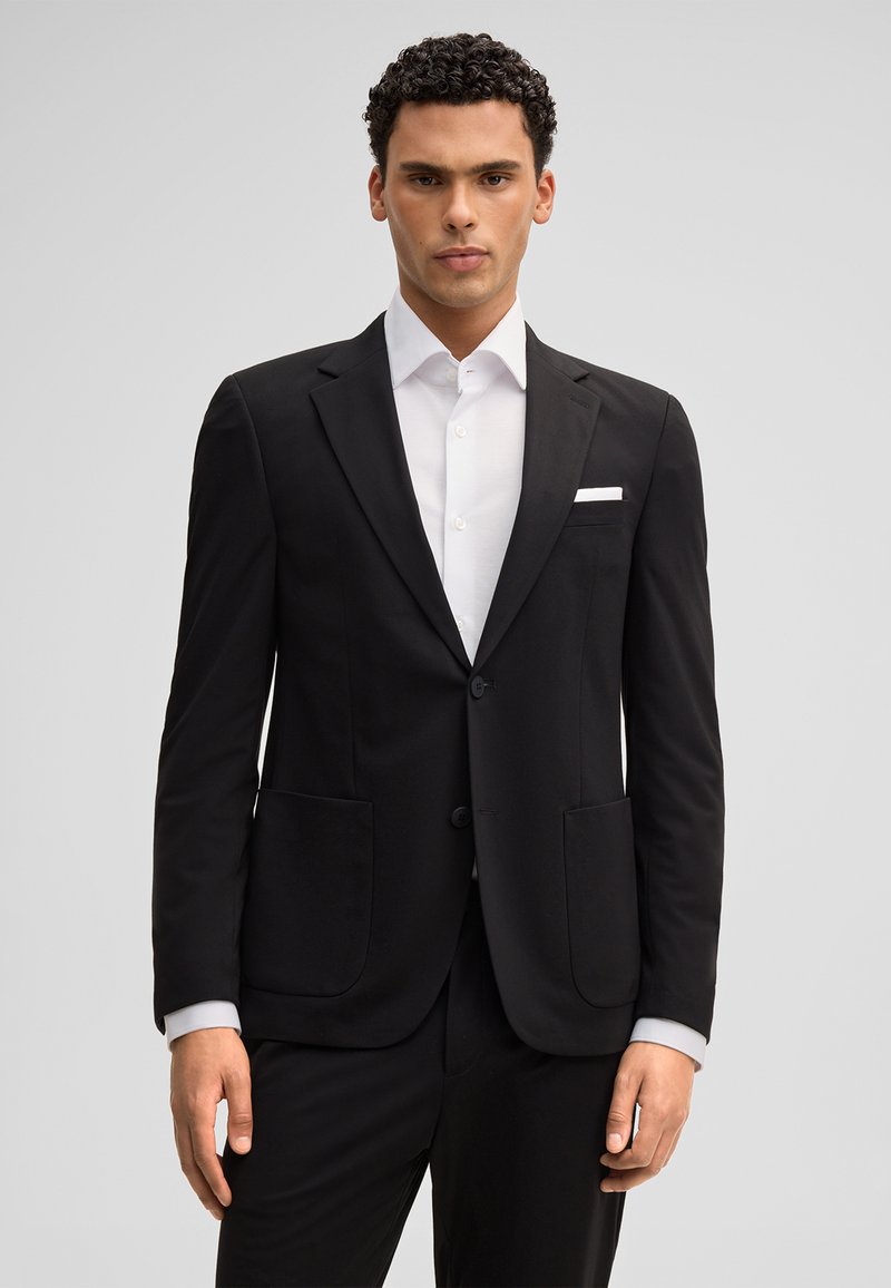 Uomo con capelli ricci che indossa una giacca da abito nera, una camicia bianca e un fazzoletto da taschino, in piedi davanti a uno sfondo grigio uniforme.