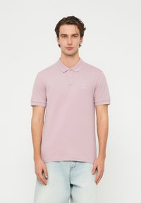 Νεαρός άνδρας στέκεται φορώντας ένα ανοιχτό μοβ πόλο πουκάμισο και ανοιχτό γαλάζιο τζιν τζιν παντελόνι σε λευκό φόντο.