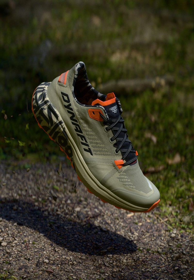 Χακί αθλητικά παπούτσια με πορτοκαλί λεπτομέρειες, επάνω μέρος από αναπνεύσιμο δίχτυ, μαύρες κορδόνες και σόλα με ανθεκτικό σχέδιο, suspendida πάνω από μια φυσική επιφάνεια.