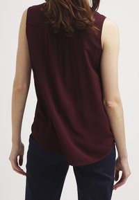 Burgundy sleeveless blouse featuring a round neckline, soft fabric, pleat details on the back, and a slightly curved hem.