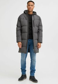 Grå polstret jakke med hætte, lynlås foran og brystlomme, båret over en sort sweater og blå jeans, kombineret med sorte støvler.