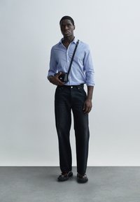 Μπλε και λευκό ριγέ πουκάμισο, σκούρο τζιν και μαύρη τσάντα χιαστί. Το μοντέλο φοράει μαύρα παπούτσια και στέκεται μπροστά σε ανοιχτό φόντο.