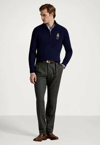 Man i marinblå kvartdrag sweater med björnlogg, grå veckade byxor, brun bälte och skor, som tittar ner med en hand i fickan.