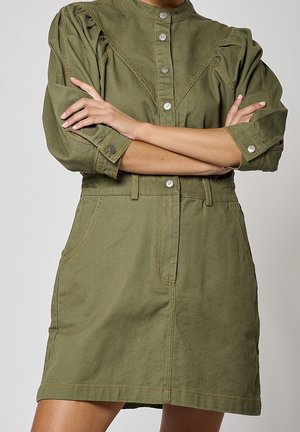 Woman wearing an olive green, buttoned dress with puffed sleeves, standing with arms crossed against a plain background.