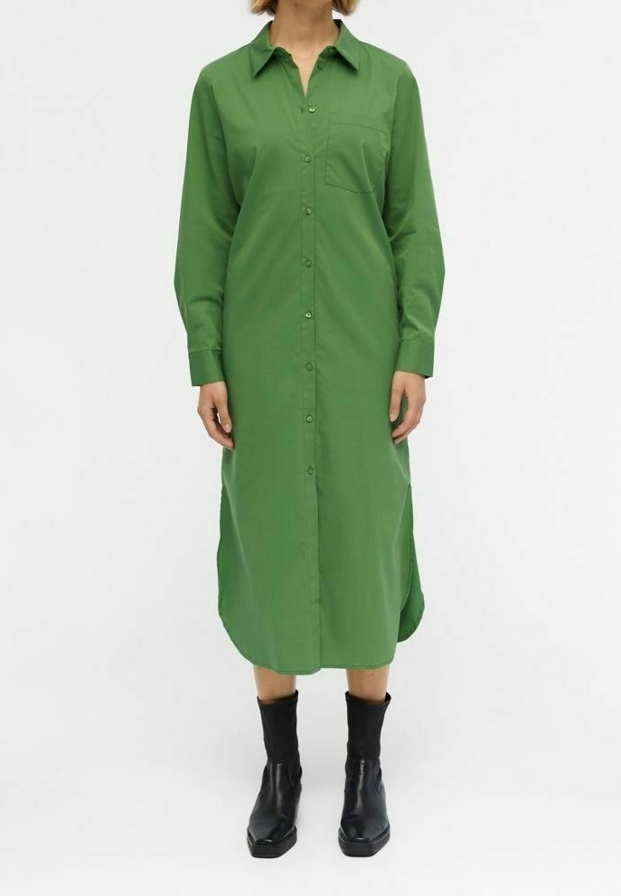 Femme portant une robe chemise midi verte à manches longues, boutonnée, avec une poche poitrine, associée à des bottes noires mi-mollet, debout devant un fond blanc.
