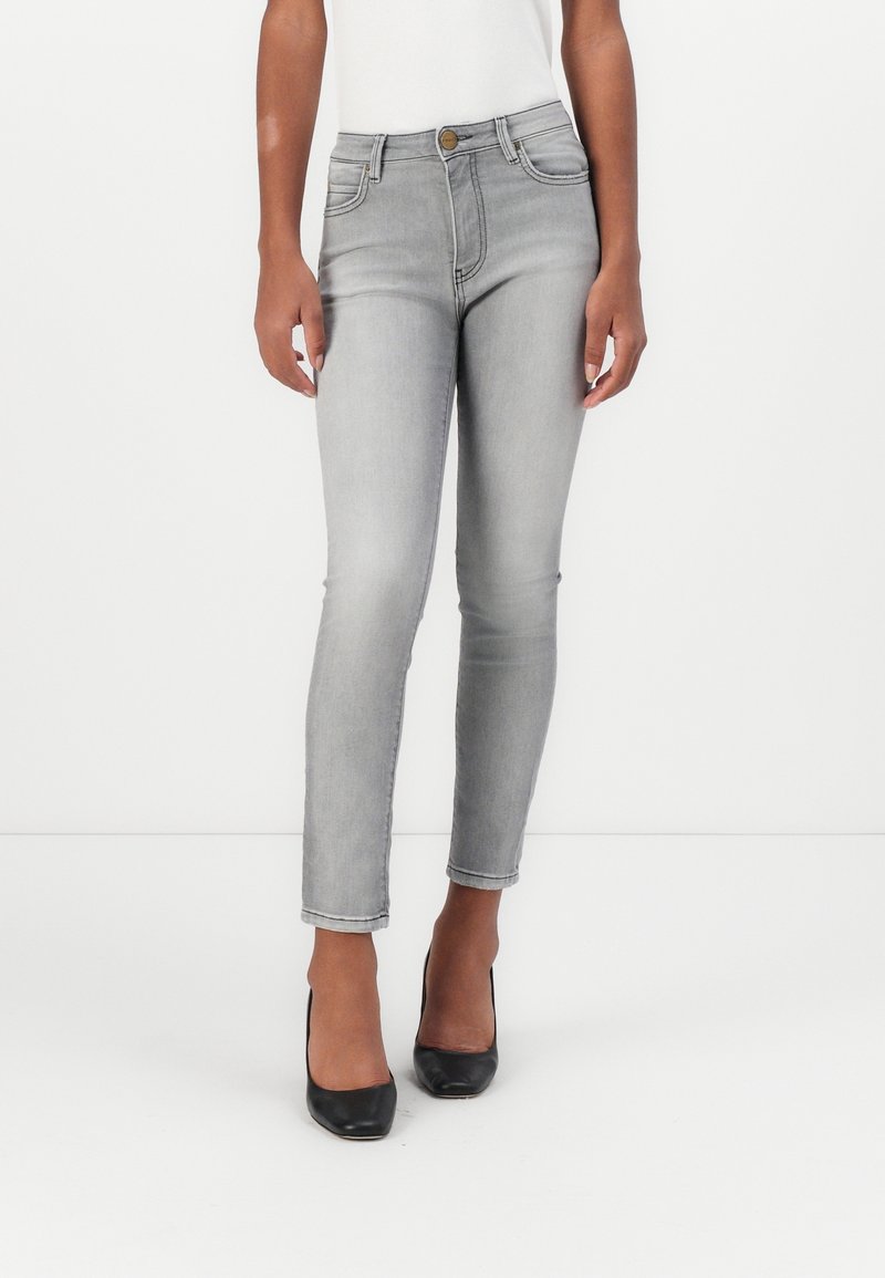 Personne portant un jean skinny gris clair et des talons noirs, debout devant un fond blanc uni, montrée de la taille aux pieds.