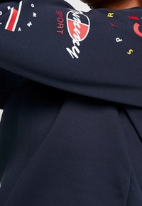 Camisa de manga larga azul marino hecha de tela suave, con impresiones de texto y logotipos multicolores en la manga. Diseño simple y contemporáneo.