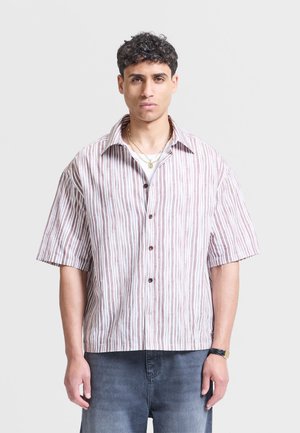 Mann med kort krøllete hår iført en kortermet, brun- og hvitstripet skjorte med knapper og mørke jeans, stående mot en ensfarget bakgrunn.