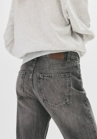 Črne jeans hlače iz denima z zbledelim izgledom, petimi žepi in rjavim usnjevim označevalcem na pasu. Oblečene z svetlo sivo svetrom.