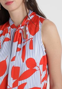 Bez rękawów bluzka w niebiesko-białym paskowanym wzorze z dużymi czerwonymi kwiatowymi motywami. Kołnierzyk z detalem w postaci zawiązanego sznurka.