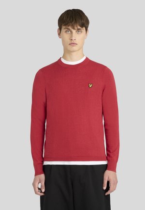 Jeune homme faisant face à l’avant portant un pull rouge à col rond avec un petit logo oiseau jaune sur la poitrine et un pantalon noir.