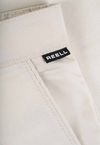 Beige cotton fabric with visible stitching, featuring a black REELL label on the seam. The texture is smooth with a durable appearance.