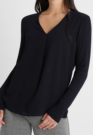 Vrouw die een zwarte blouse met lange mouwen en V-hals draagt, met grijze broek met visgraatmotief, staand met handen bij de heupen.