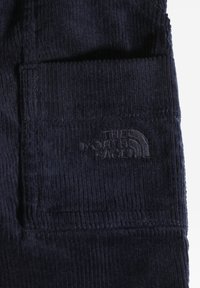 Tessuto in corduroy di un blu navy scuro con righe verticali. Presenta una tasca laterale cucita e un logo embossé nell'angolo. Texture morbida.