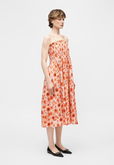 Γυναίκα που στέκεται φοράει ένα πορτοκαλί φόρεμα μέσης μήκους με λουλούδια χωρίς μανίκια, με σφηκωμένο μπούστο και μαύρα μυτερά παντόφλες, σε απλό φόντο.