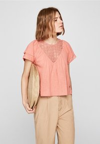 Top a maniche corte di colore rosa cipria con colletto in crochet testurizzato, abbinato a pantaloni beige e una borsa rotonda in paglia.