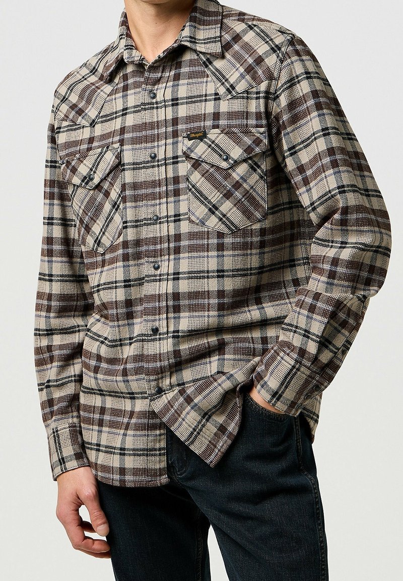 Homme portant une chemise à carreaux marron et beige à boutons avec poches rabat, associée à un jean foncé, debout devant un fond uni.