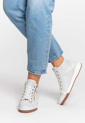 Person wearing light blue jeans and white high-top sneakers with gold eyelets and side zipper, standing on white background.