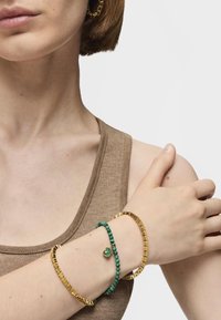 Pulsera de oro con diseño texturizado, pulsera de cuentas verdes con un charm, llevada en una muñeca de tono de piel clara, sobre un fondo neutral.