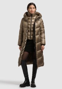 Polstret, lang olivengrønbrun frakke med hætte, der har lynlås, sidelommer og en glat, skinnende tekstur; lagdelt over en kort vest.