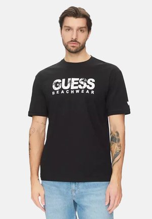 Uomo con maglietta nera "GUESS BEACHWEAR" e jeans azzurri, in piedi con tatuaggi visibili su entrambe le braccia.