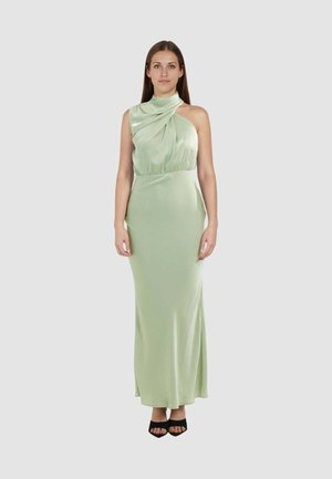 Donna in piedi con un abito da sera senza maniche in raso verde chiaro con scollo alto asimmetrico, indossa tacchi neri aperti sulle dita.