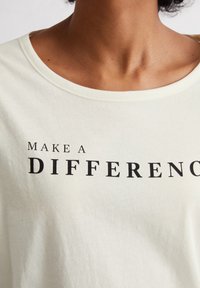 T-shirt en coton crème clair avec un col rond et un texte noir qui dit "FAITES UNE DIFFÉRENCE" affiché en évidence sur le devant.