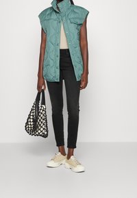 Chaleco sin mangas de color azul verdoso acolchado con cierres a presión, pantalones ajustados negros, zapatillas gruesas beiges y un bolso tejido blanco y negro.