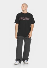Czarna bawełniana koszulka z odważnym graficznym napisem "PROSTO" w gradientowych odcieniach, zestawiona z ciemnoszarymi spodniami cargo z wieloma kieszeniami i luźnym krojem.